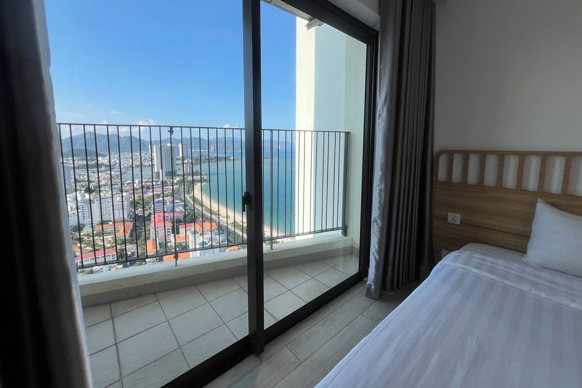 A hotel room with a large balcony, a bed, and a view of a coastal cityscape.