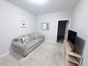 Un salon avec un canapé gris, un téléviseur à écran plat posé sur un meuble en bois, et une porte donnant sur une autre pièce.