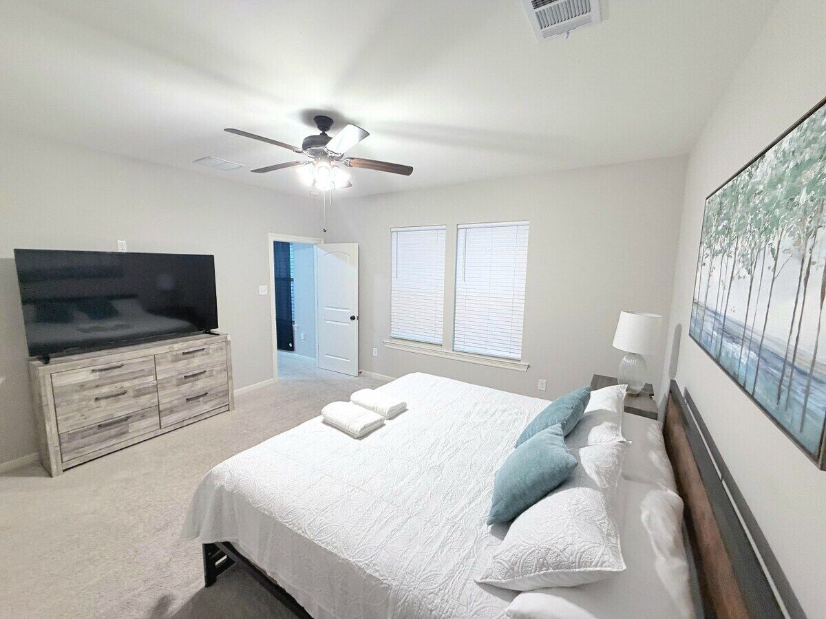 A bedroom with a bed, a TV, a ceiling fan, and a door leading to another room.