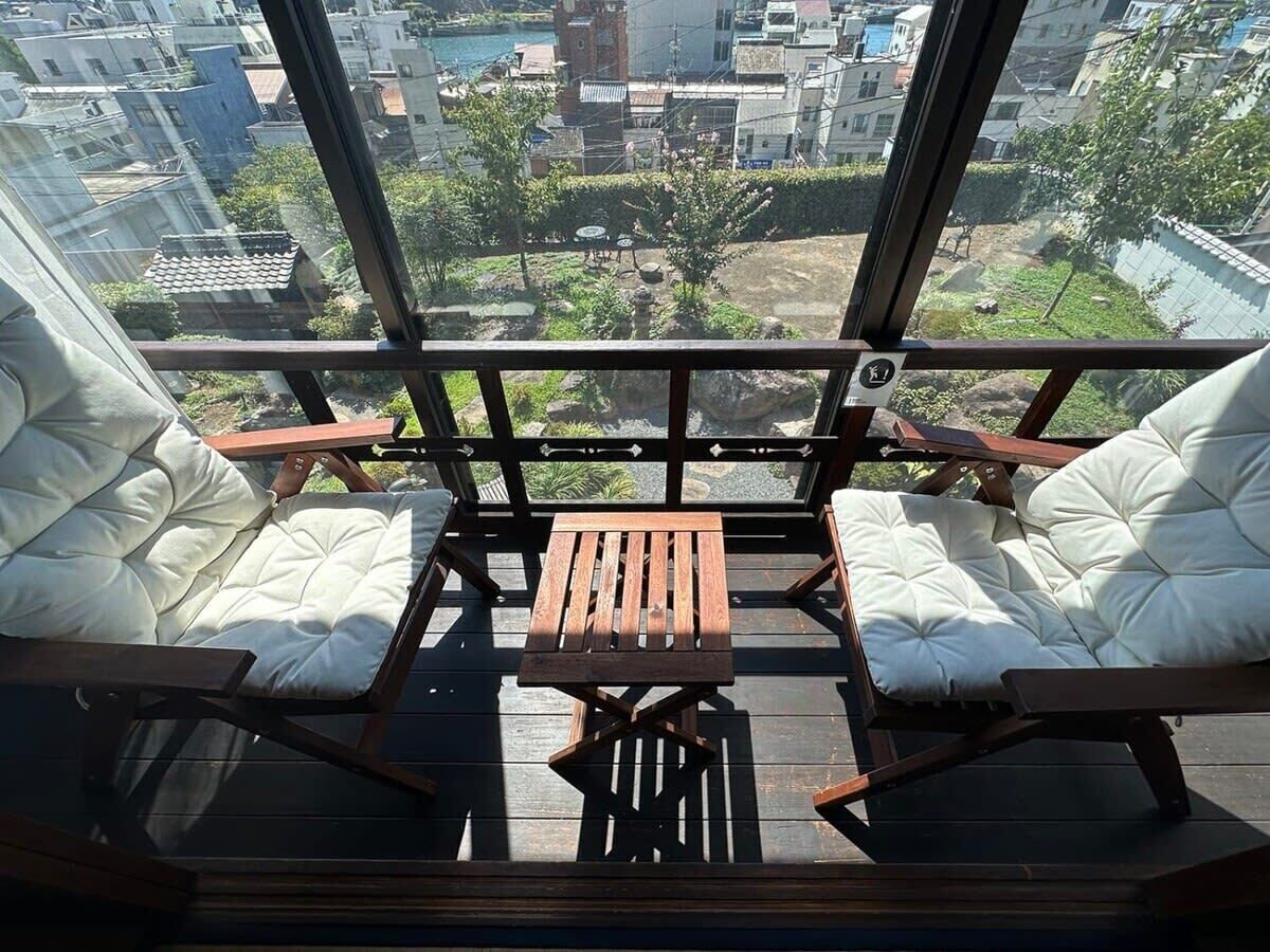 A balcony with two cushioned chairs and a wooden table, overlooking a cityscape with buildings and greenery.
