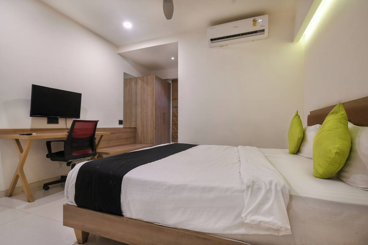A hotel room with a bed, a desk with a chair, a television, and an air conditioning unit.