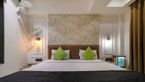 Quarto de hotel com uma cama grande, duas almofadas verdes, uma televisão de tela plana e um abajur fixado na parede.