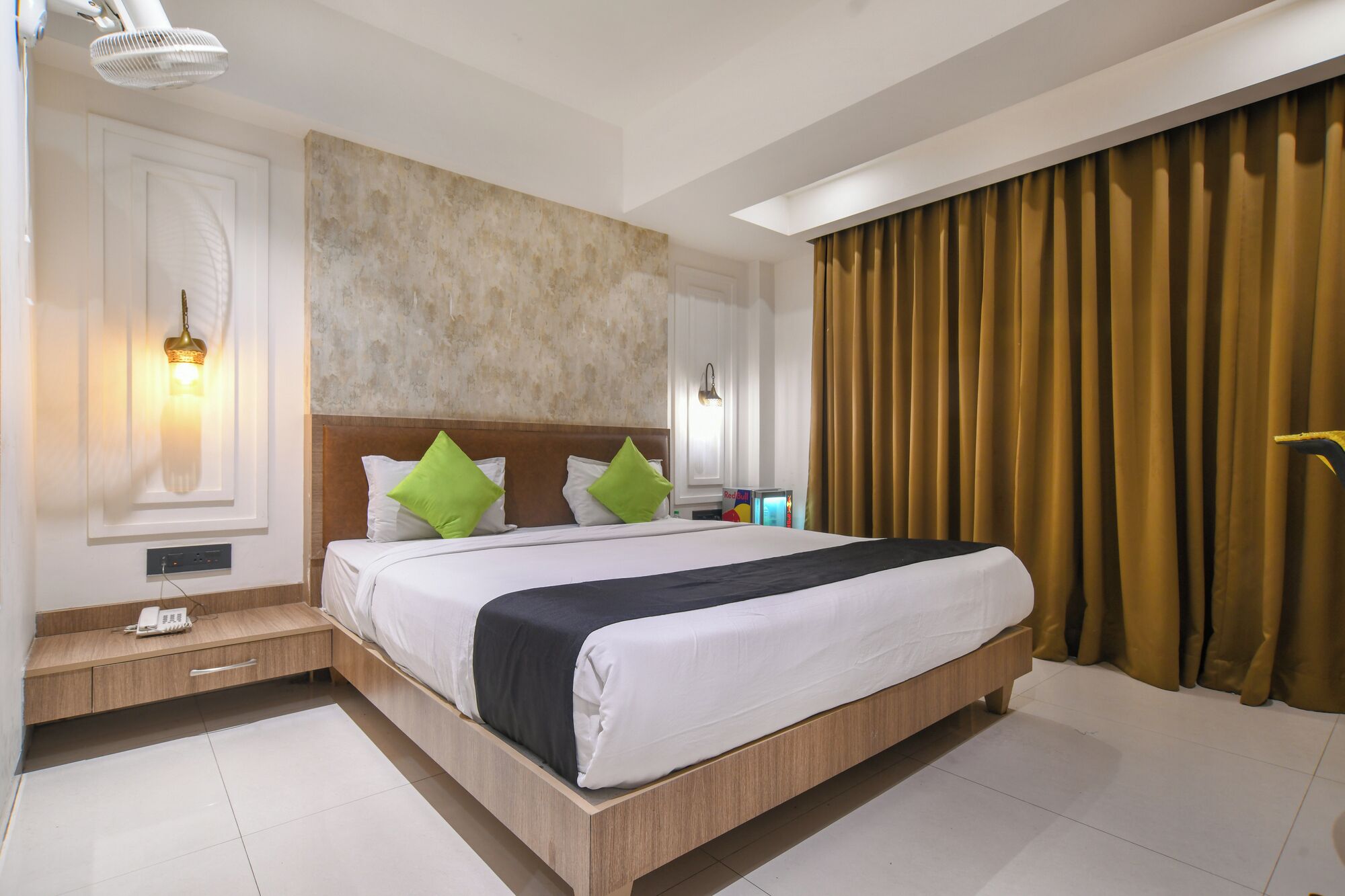 Una habitación de hotel con una cama grande, dos lámparas de noche, una mesita de noche, una luz en la pared y una ventana con cortinas.