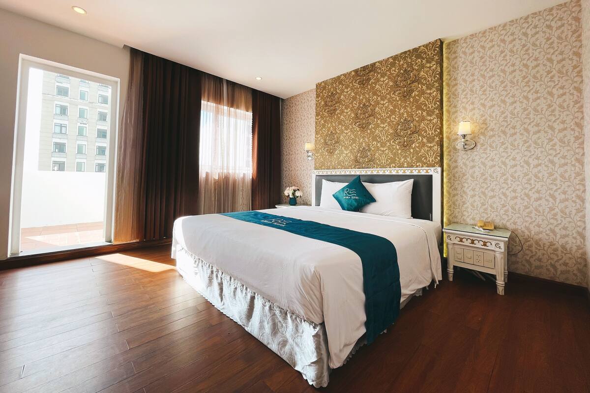 A hotel room with a large bed, a nightstand, a wall-mounted lamp, and a view of a building through the window.
