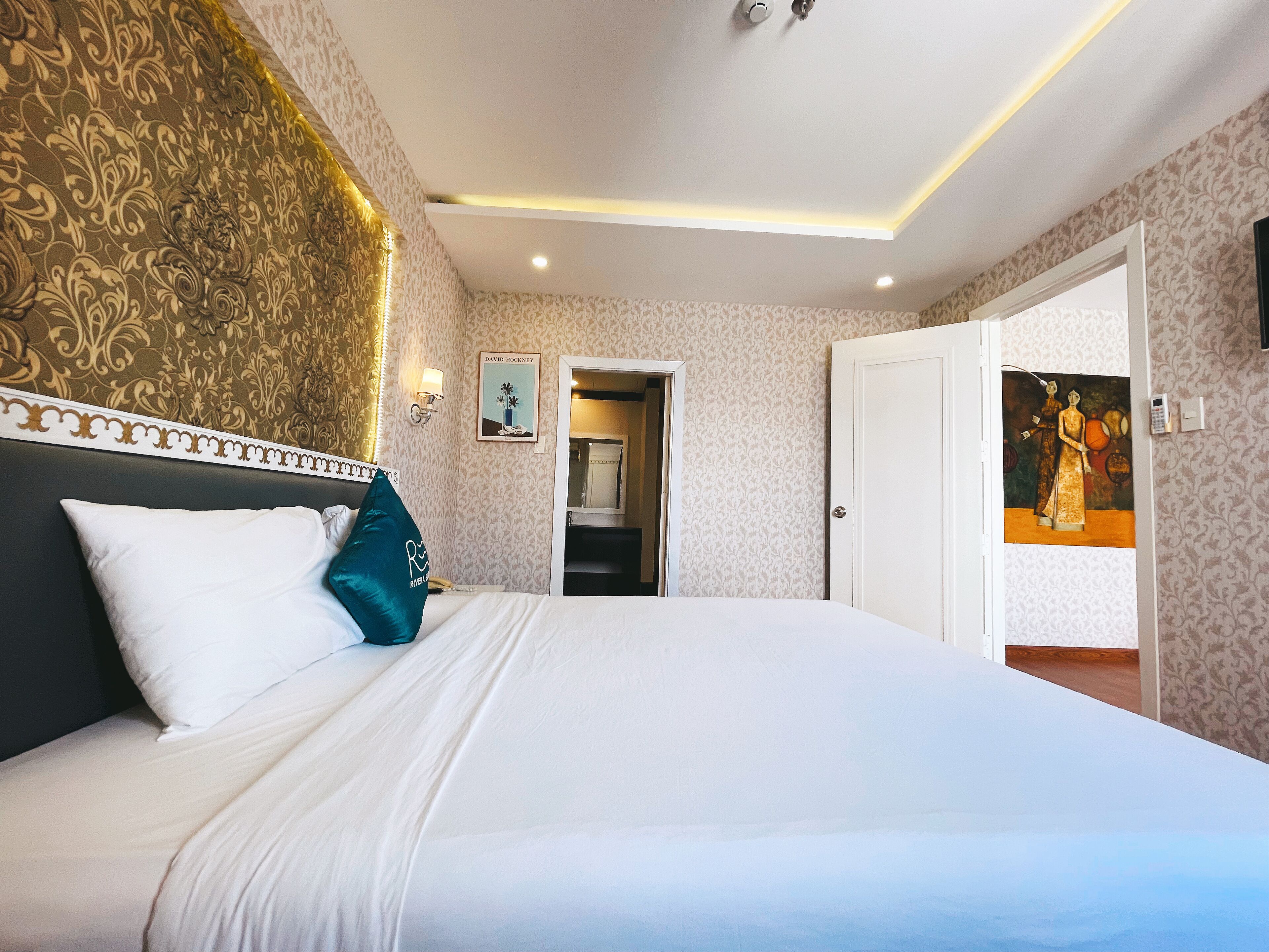 A hotel room with a large bed, a headboard with a decorative pattern, a painting on the wall, and a door leading to another room.