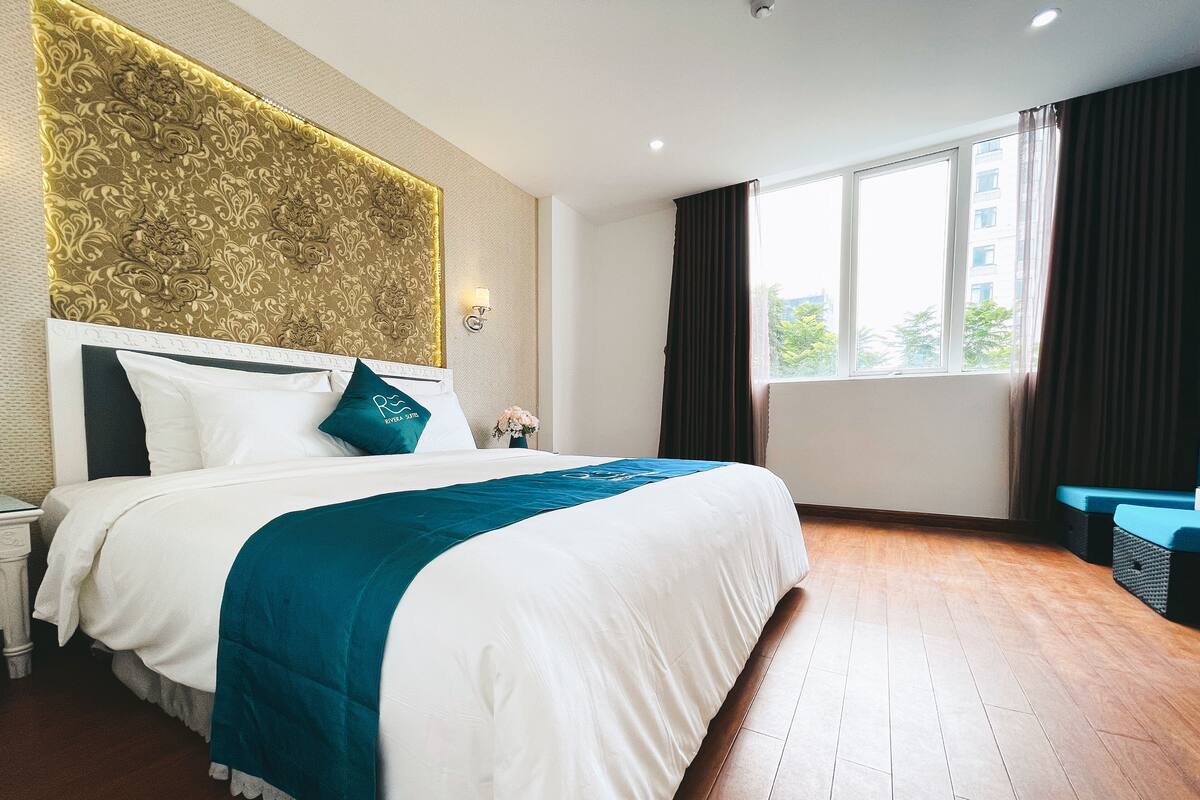 A hotel room with a large bed, a nightstand, a wall-mounted lamp, a window with curtains, and a view of the outside.