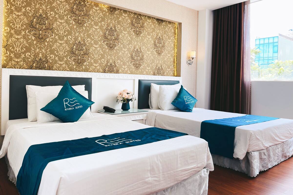 A hotel room with two beds, a decorative gold-patterned wall, a nightstand with a vase of flowers, and a window with curtains.