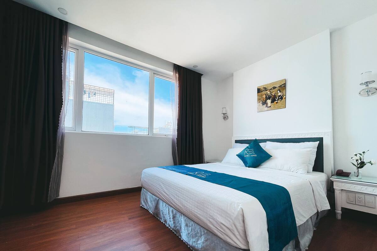 A hotel room with a large bed, a nightstand, a window with curtains, and a painting on the wall.