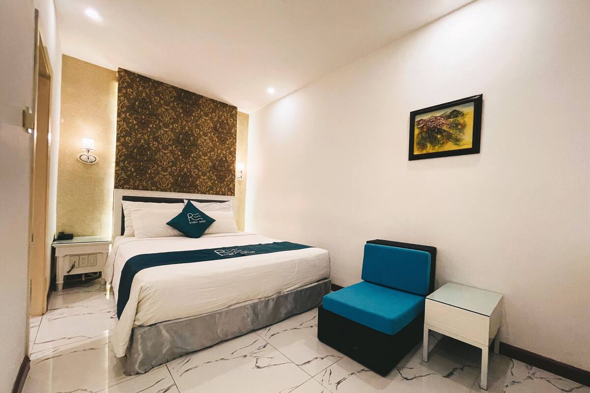 A hotel room with a bed, a bedside table, a chair, a nightstand, and a wall with a painting.