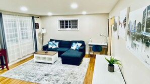 A modern living room with a dark blue sofa, a white coffee table, and a small side table. There is a window with white curtains, a wall-mounted shelf, and a framed cityscape picture.