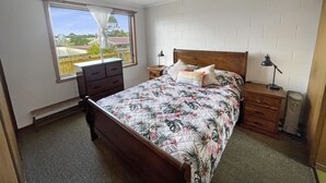 A bedroom with a bed, a dresser, a nightstand, a lamp, and a window with a view of greenery.