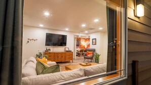 A modern living room with a sofa, a wooden cabinet, a TV, and a dining area with chairs and a table. - Mid-Mod Urban Hub | Hot Tub & Fire Pit (Port Angeles)