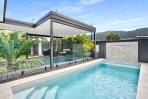 Un área de piscina moderna al aire libre con un porche acristalado, mobiliario y vistas del paisaje circundante.