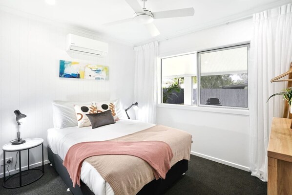 Un dormitorio con cama, mesita de noche, lámpara, ventana y ventilador de techo.