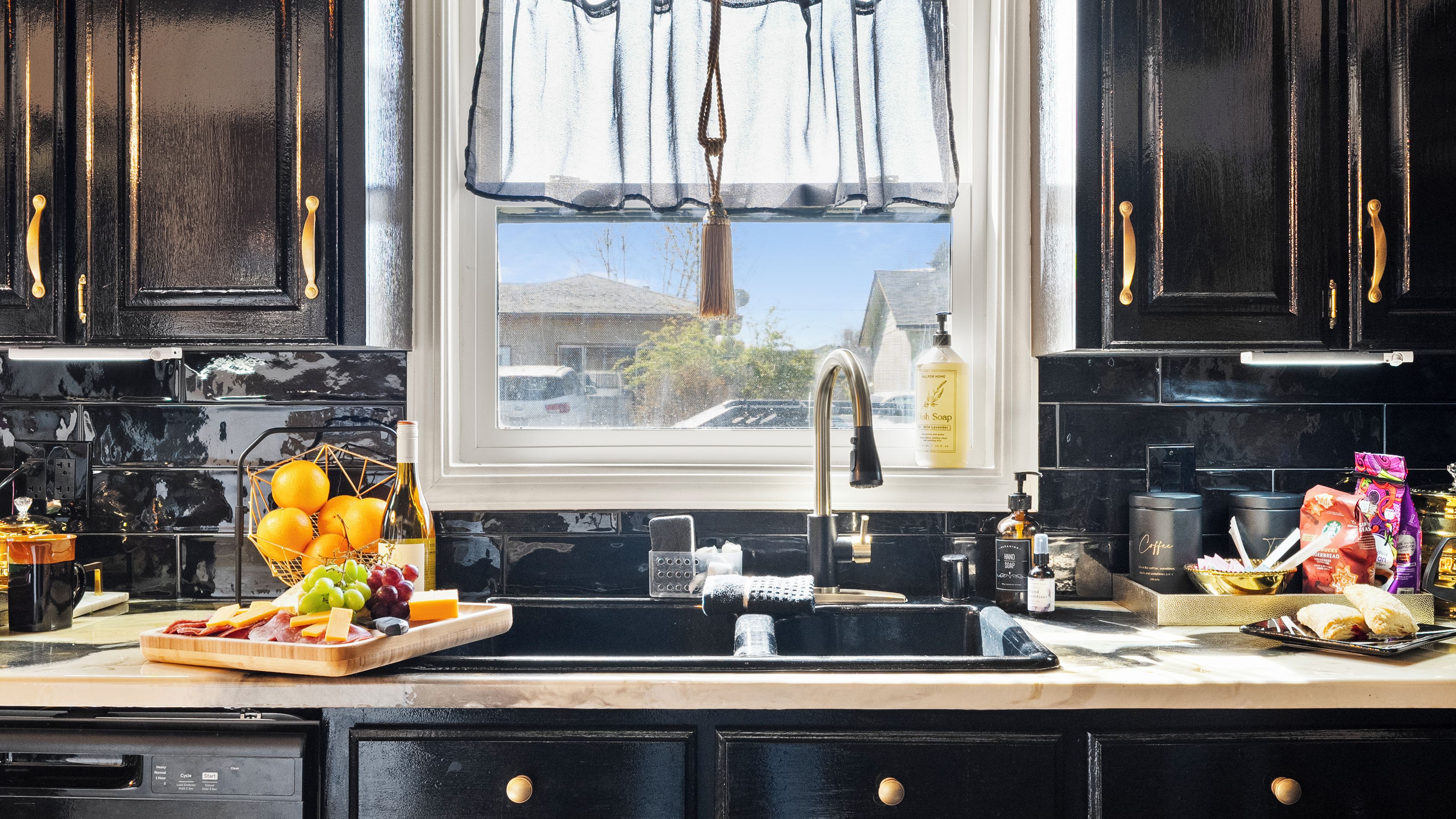 A kitchen with black cabinets, a window with a view of houses, a sink, and various kitchen items.