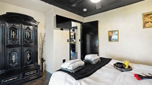A hotel room with a large black wardrobe, a bed with neatly folded towels, a tray with a cup and orange juice, and a painting on the wall. - THE ONYX — A GOTHIC WINE-COUNTRY HOME WITH A HOT TUB, FIRE PIT AND GAME ROO... (College Place)