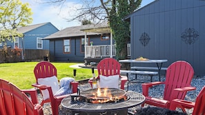 Ein Garten mit Feuerstelle, roten Adirondack-Stühlen, einem Picknicktisch und einem Haus mit Veranda.