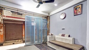 Een kamer met een gesloten luik, een blauw hek met een bord waarop staat 'Toegang Verboden', een bank, een klok en een plafondventilator.