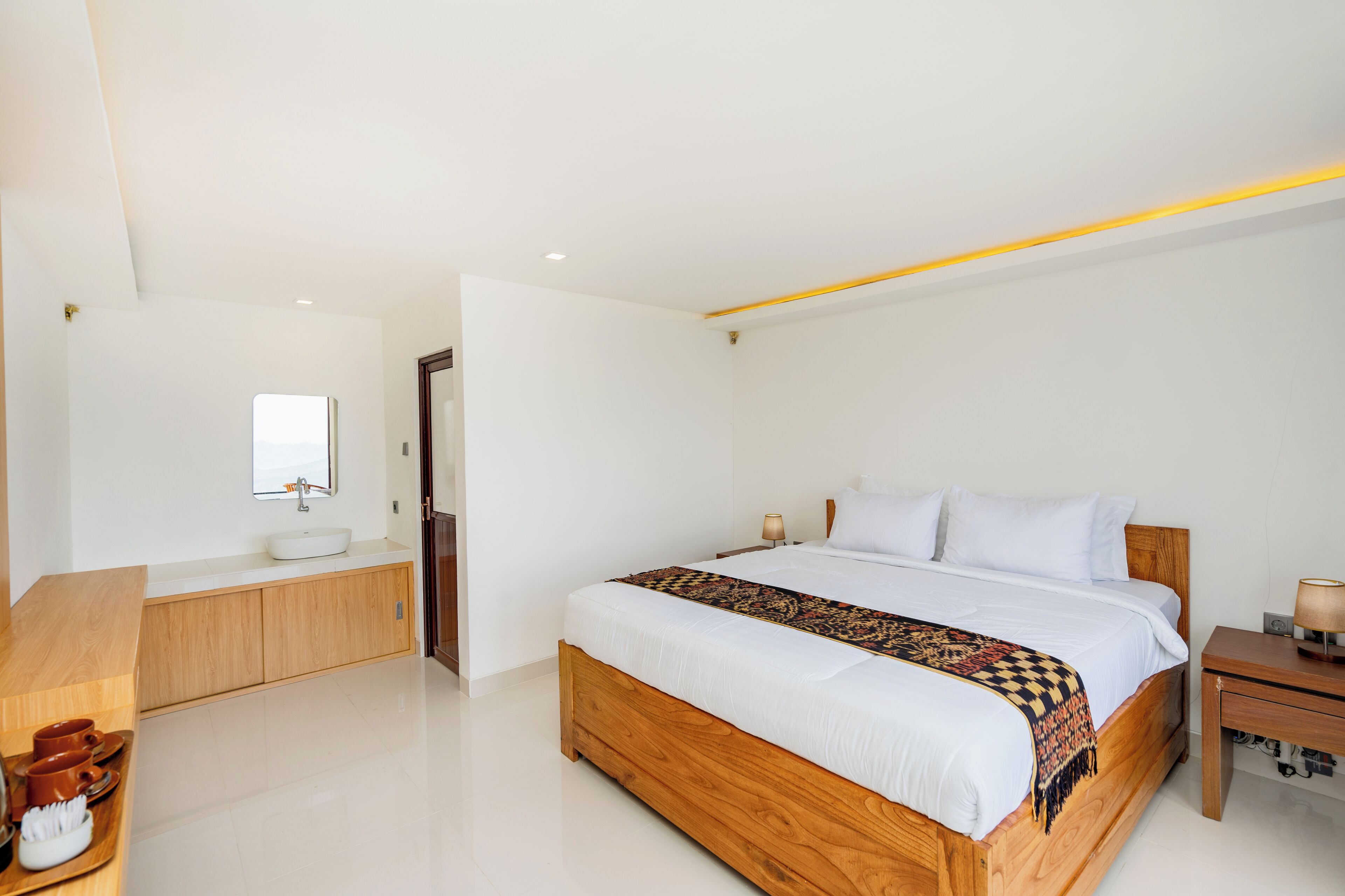 A modern bedroom with a wooden bed, white bedding, a bedside table, and a bathroom with a sink and mirror.