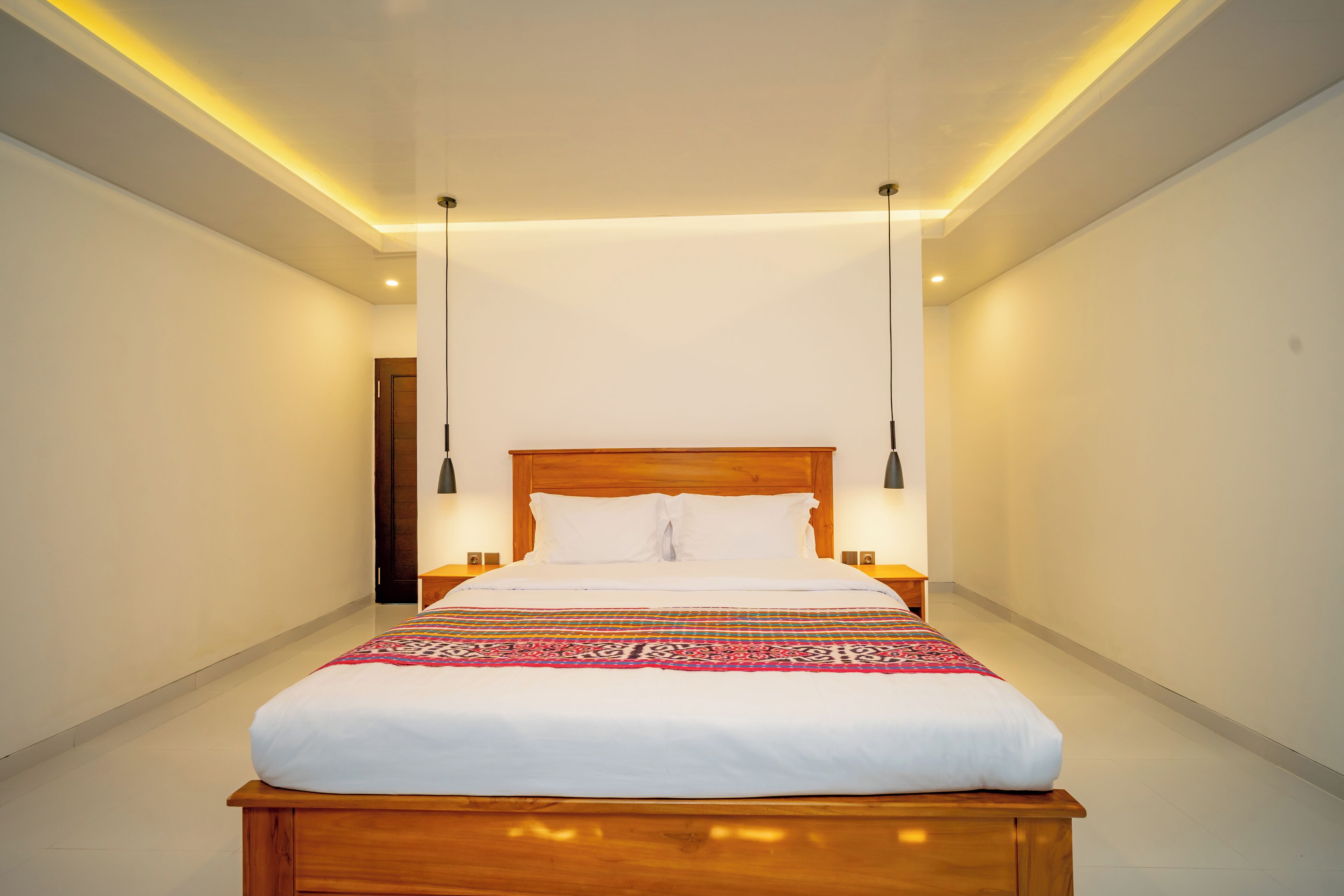 A neatly made bed with a wooden headboard and white bedding, flanked by two bedside tables with lamps, in a well-lit room with a high ceiling and a door on the left.