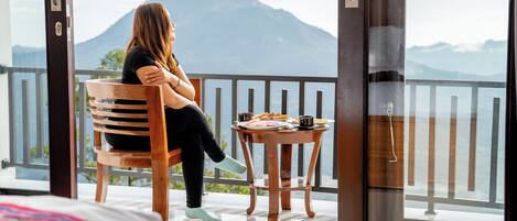Una persona sentada en una silla de madera junto a un balcón con vistas a la montaña, una mesa pequeña con comida y una cama con una manta estampada.