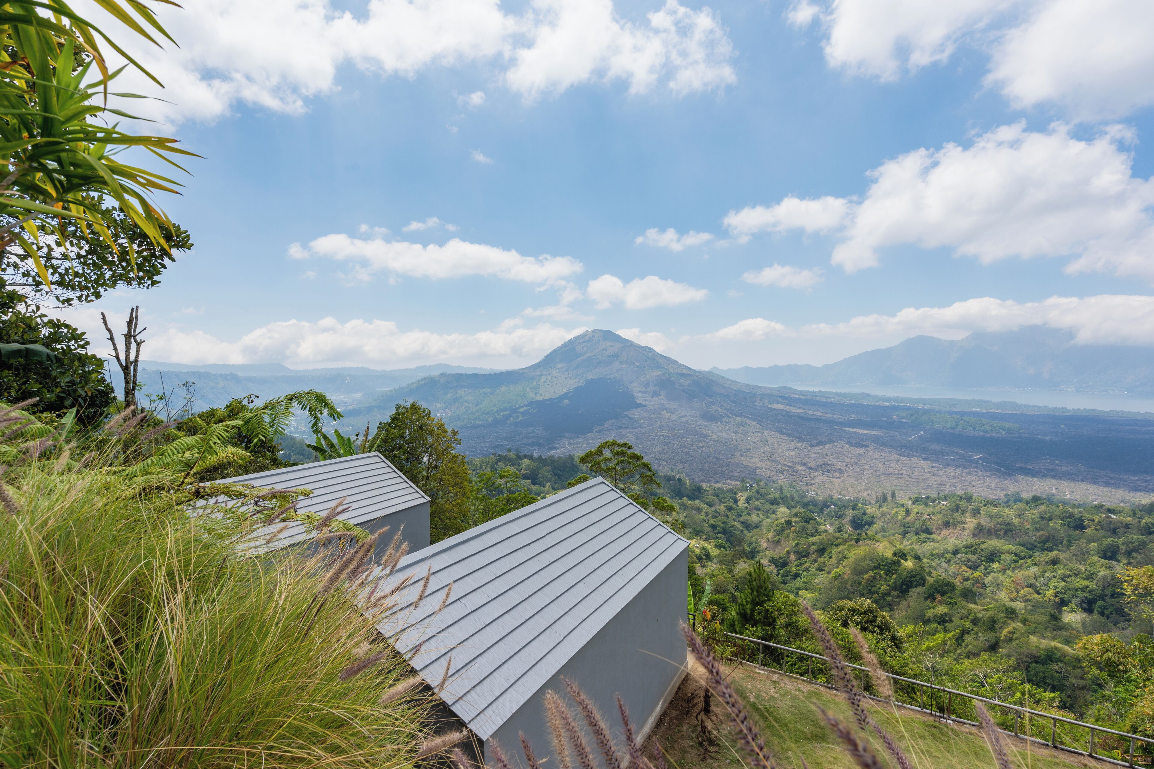 A view of a mountainous landscape with a prominent peak in the distance. There are two buildings with grey roofs in the foreground, surrounded by lush greenery.