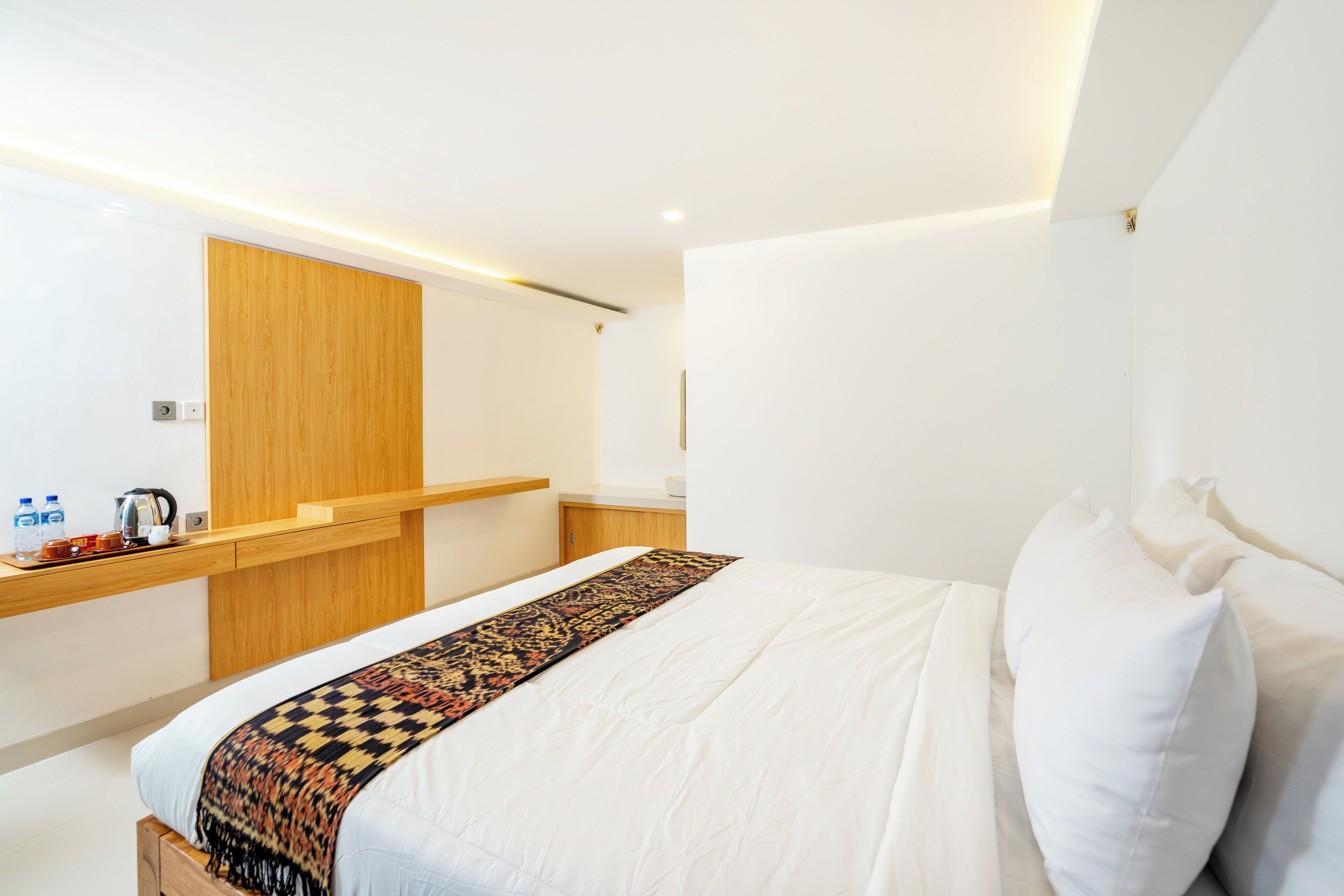 A modern hotel room with a large bed, a wooden headboard, a bedside table with a kettle, and a small shelf with toiletries.