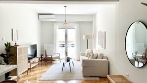 Ein modernes Wohnzimmer mit Sofa, Couchtisch und Fernseher. Ein großes Fenster mit Vorhängen, eine Hängelampe und ein Spiegel an der Wand sind zu sehen.
