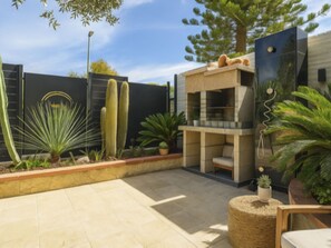 A modern outdoor patio with a stone floor, a built-in seating area, and various plants including cacti and palms. - Il Nido di Mennenne by Interhome (Villasimius)
