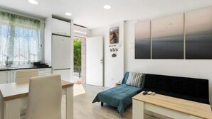 Une pièce moderne et bien éclairée, comprenant un coin cuisine, un lit recouvert d’une couverture verte, un bureau avec une chaise et une grande fenêtre ornée de rideaux légers.