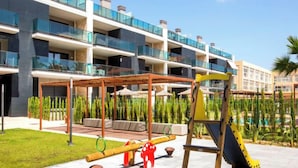 A playground with a seesaw, a slide, and a climbing structure in front of a modern apartment building with balconies. - Golden Ray by Interhome (Jávea)