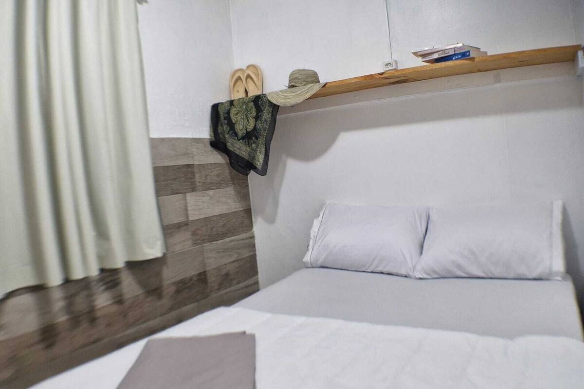 A hotel room with a bed, a wooden shelf above it, a pair of slippers, and a folded item on the bed.