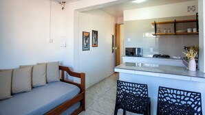 Un espace de vie compact comprenant un canapé, un coin repas avec des chaises, une cuisine avec un plan de travail et une porte donnant sur une autre pièce.