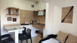 Une petite cuisine équipée d’un four à micro-ondes, d’une bouilloire et d’un petit évier. On y trouve deux chaises noires et un lit avec des oreillers de couleur beige.