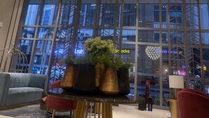 A modern lobby with a large spherical chandelier, a seating area with a blue sofa, and a view of a cityscape through large windows.