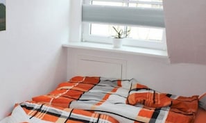 A small, neatly made bed with a checkered blanket in a room with white walls and a window with blinds.