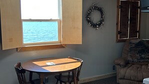 A room with a round wooden table and chairs, a window with a view of the sea, a wreath on the wall, and a brown armchair. - Small Space with a BIG View!!! (Redfield)