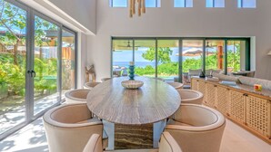 Un comedor espacioso con una mesa redonda y sillas, ventanas amplias con vistas al océano y un balcón con mobiliario exterior.