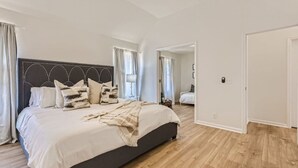 A bedroom with a large bed, a dark headboard, and a wooden floor. There is a door leading to another room.