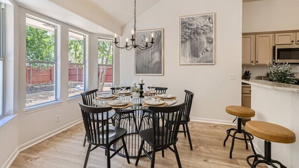 A dining area with a round table, black chairs, and a chandelier. There are windows with a view of trees and a red fence.