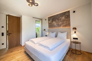 Quarto moderno com cama grande, porta de madeira, vista para área verde e luminária fixada na parede.