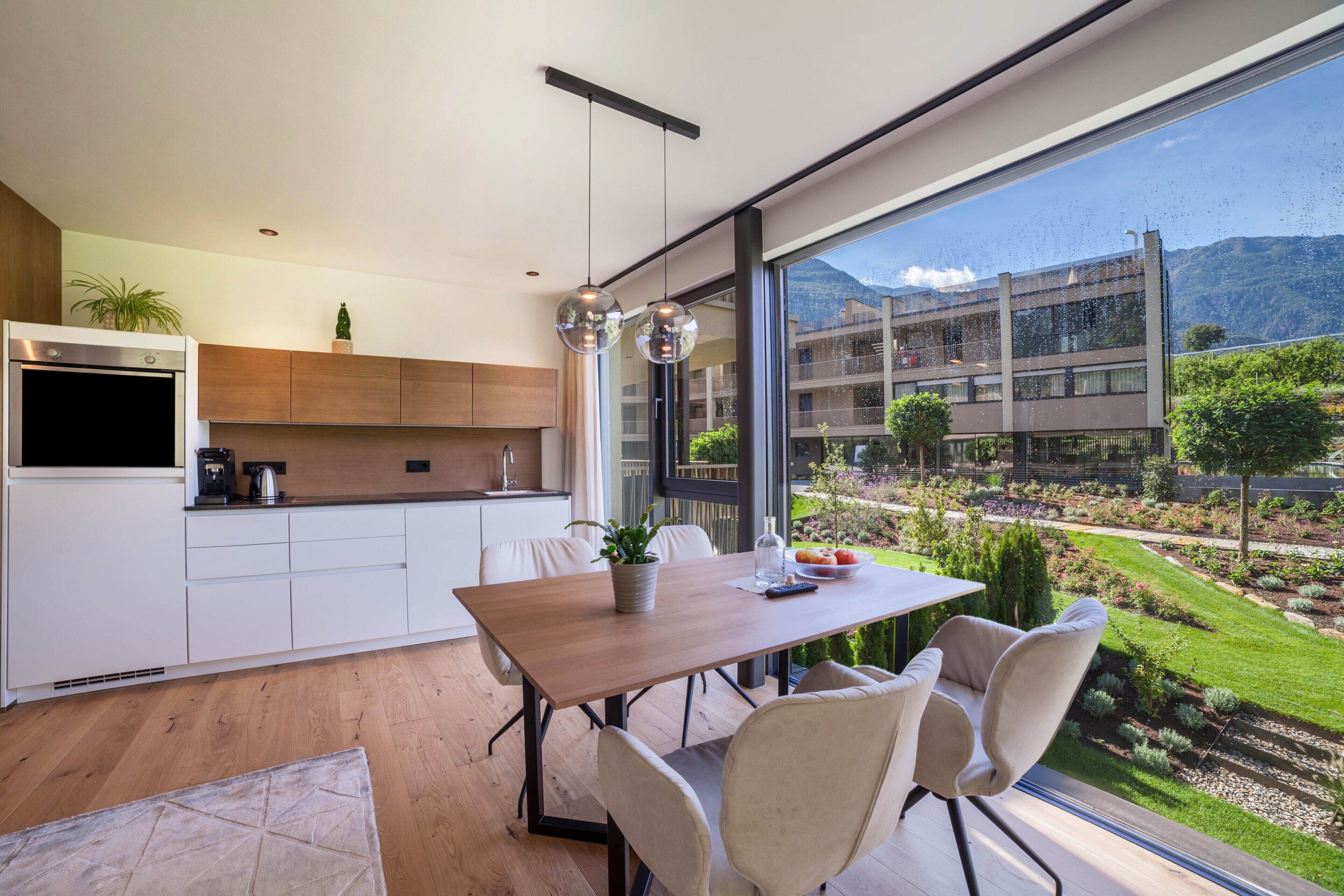 A modern kitchen with a dining area, large windows, and a view of a building and garden.