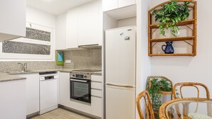 A modern kitchen with white cabinets, granite countertops, a built-in oven, a refrigerator, and a dining area with wicker chairs and a glass table. - Holiday Apartment "Aubi" with Open Terrace and Indoor Terrace (Palamós)