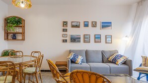 A living room with a glass coffee table, wicker chairs, a grey sofa with patterned cushions, a wall-mounted plant shelf, and framed artwork. - Holiday Apartment "Aubi" with Open Terrace and Indoor Terrace (Palamós)
