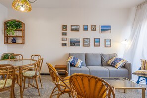 A living room with a glass coffee table, wicker chairs, a grey sofa with patterned cushions, a wall-mounted plant shelf, and framed artwork.