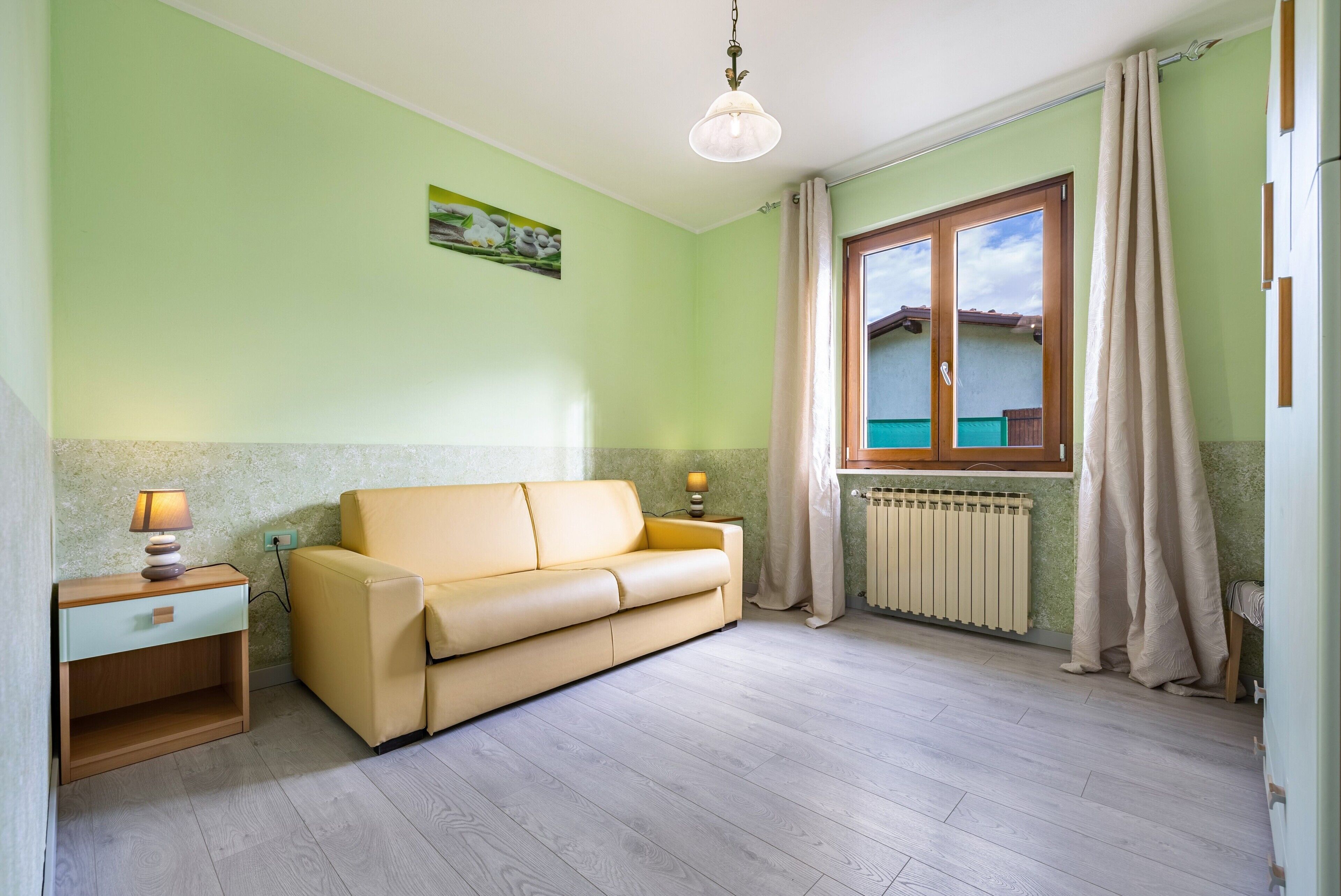 Une pièce avec un canapé beige, une petite table de chevet en bois avec une lampe, un radiateur et une fenêtre avec des rideaux.