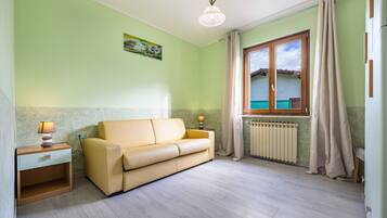 Une pièce avec un canapé beige, une petite table de chevet en bois avec une lampe, un radiateur et une fenêtre avec des rideaux.