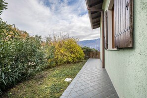 A pathway leading to a building with a green exterior, wooden shutters, and a view of mountains.