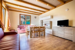 A cozy living room with a sofa, a dining table with chairs, a TV on a cabinet, and a window with a view of a house and greenery.