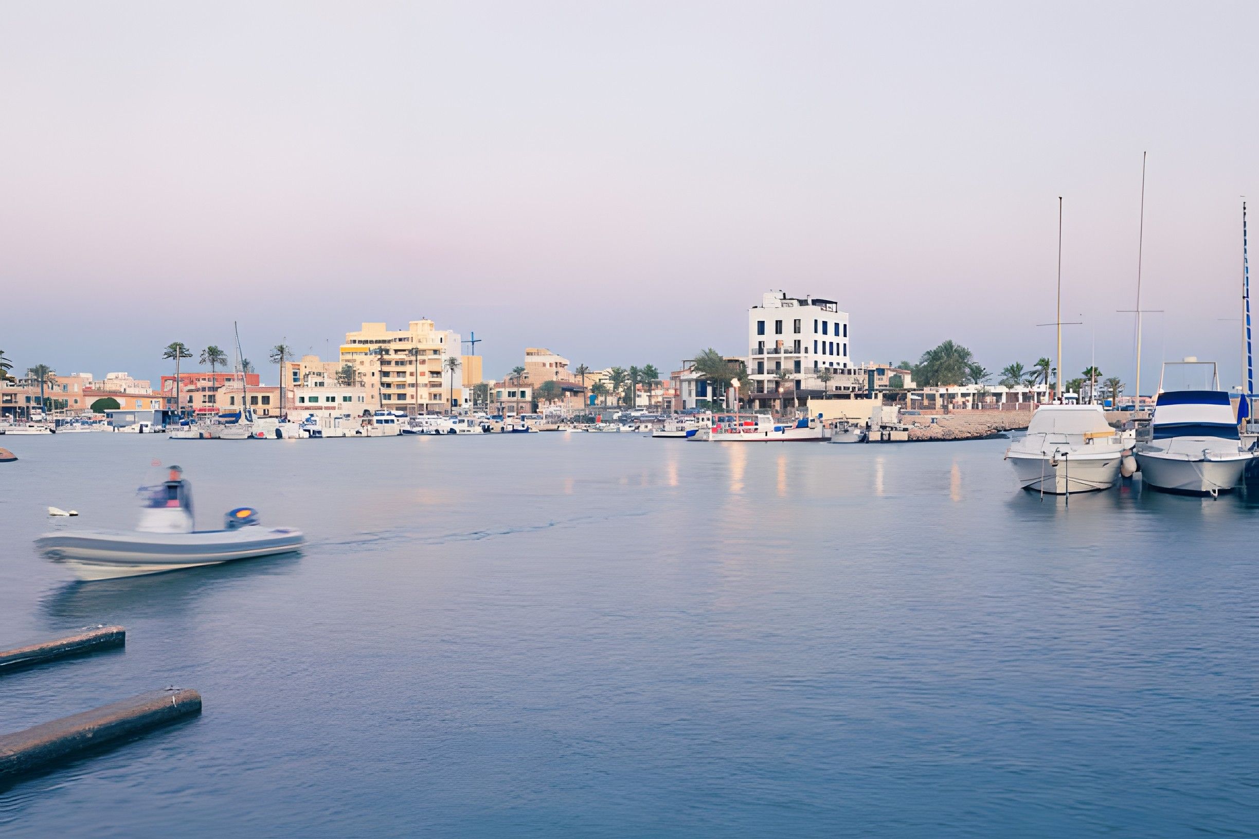 Un port de plaisance avec des bateaux amarrés, une étendue d’eau calme et un paysage urbain avec des bâtiments en arrière-plan.